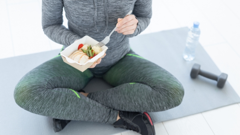 Osoba u sportskoj opremi sedi na podlozi i jede lagan obrok pre treninga, pored bučice i flaše vode